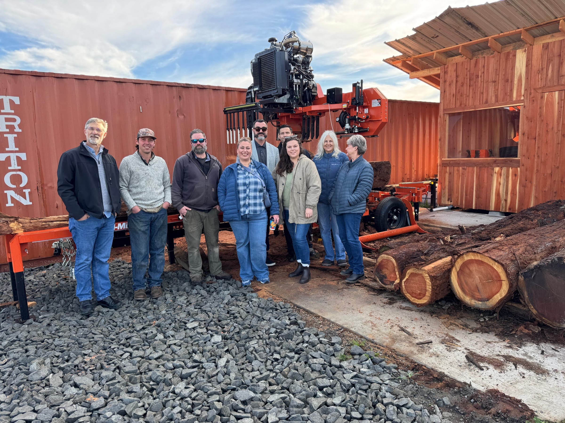 Nine members of the BLREDCO team standing together in front of the Tribe's portable sawmill, with freshly milled redwood logs stacked to one side.